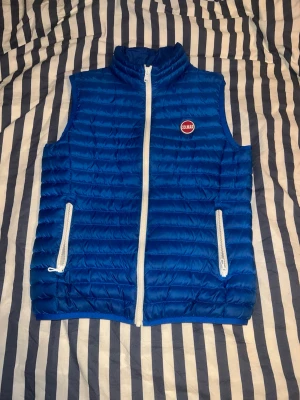 Blå dunväst från Colmar - Snygg blå dunväst från Colmar med vit dragkedja och två praktiska fickor framtill. Västen har quiltad design och står ut med sitt klassiska Colmar-märke på bröstet. Insidan är ljusgrå och materialet är lätt och smidigt, perfekt för lager-på-lager. En skön väst att ha på sommarn och ett riktigt bra pris. Denna väst passar in mer i grisch stilen men går att ha på sig till alla sorters kläder. PRIS kan diskuteras. Fraktar på 1-2 dagar!!!!