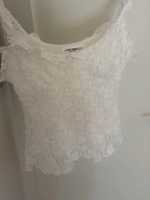 Vit spetslinnetopp från Pull&Bear - Säljer en söt vit linnetopp från Pull&Bear med spetsdetaljer och stretchig struktur. Toppen är ärmlös med breda axelband och har en lätt croppad passform. Perfekt för dig som gillar romantisk och trendig stil.