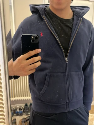 Mörkblå hoodie från Ralph Lauren - Säljer en mörkblå hoodie med dragkedja från Ralph Lauren. Klassisk design med huva, två fickor framtill och den ikoniska röda loggan broderad på bröstet. Perfekt för en avslappnad och stilren look. Står att det är xl men de är i barnstorlek skulle jag gissa. Pasar som en S!