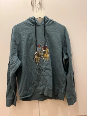 Blå hoodie från U.S. Polo Assn - Snygg blå hoodie från U.S. Polo Assn med broderat motiv av två polospelare på hästar framtill. Tröjan har huva med dragsko och ribbade muddar. Perfekt för en avslappnad och sportig stil.