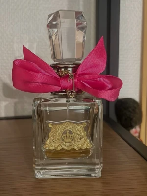 Juicy Couture parfym med rosa rosett - Juicy Couture parfym i en lyxig, fyrkantig glasflaska med en stor, kristalliknande kork och en dekorativ rosa rosett runt halsen. Flaskan har en guldfärgad emblem på framsidan som ger en exklusiv känsla. Perfekt för dig som gillar snygga detaljer på parfymhyllan.