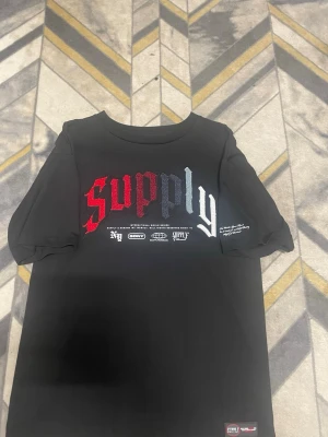 Svart t-shirt Supply & Demand - Cool svart t-shirt från Supply & Demand med broderad text 'supply' i rött, grått och vitt framtill. Rund halsringning och tryck med loggor under texten. Perfekt för en streetwear-look.