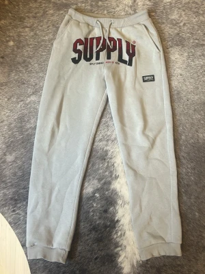 Gråa mjukisbyxor från SUPPLY - Snygga gråa mjukisbyxor från SUPPLY med stort tryck framtill och dragsko i midjan. Byxorna har ribbade muddar vid bensluten och två sidofickor. Perfekta för en chill och avslappnad stil. Materialet känns mjukt och bekvämt, troligen bomullsmix.