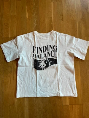 Vit t-shirt Finding Balance 157. - Vit t-shirt från 157. med trycket 'FINDING BALANCE' och ett svartvitt yin-yang-motiv på bröstet. Klassisk passform med rund hals och korta ärmar. Tillverkad i 100% bomull, perfekt för en avslappnad stil.
