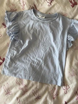 Ljusblå topp med volangärmar KappAhl - Supersöt ljusblå topp från KappAhl med korta volangärmar. Enkel och stilren design i mjuk bomull, perfekt för en fräsch och trendig look. Passar till både jeans och kjol för en chill vibe.