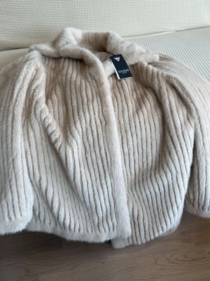 Beige pälsjacka från Guess - Snygg beige pälsjacka från Guess med mjuk fuskpäls och diskret randigt mönster. Jackan har klassisk krage och helfodrad insida med Guess-logga. Perfekt för att hålla värmen och samtidigt se trendig ut under vintern.