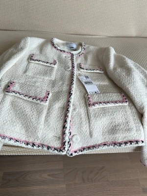 Vit cardiganjacka med rosa detaljer - Stilren vit cardiganjacka från mango i bouclé-liknande material. Jackan har fyra fickor framtill och dekorativa kanter i rosa, svart och vitt. Rund halsringning och knäppning framtill. Perfekt för en trendig och chic look.