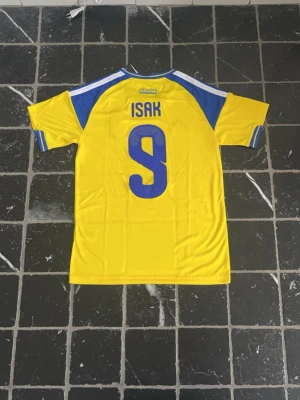 Sverige fotbollströja Isak #9 Adidas - Gul och blå Sverige fotbollströja från Adidas med Isak och nummer 9 på ryggen. Tröjan har klassiska Adidas-ränder på axlarna, broderad landslagssköld och är tillverkad i lätt funktionsmaterial. Perfekt för match eller träning.