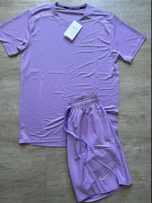 Lila träningsset från Nike - Säljer ett lila träningsset från Nike med t-shirt och shorts. Setet är gjort i ett lätt och snabbtorkande material, perfekt för träning. T-shirten har rund hals och korta ärmar, shortsen har resår och dragsko i midjan. Diskret Nike-logga på båda plaggen.