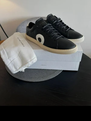 Svarta sneakers från Oliver Cabell - Stilrena svarta sneakers från Oliver Cabell med vit sula och en stor vit cirkeldetalj på sidan. Skorna har svarta skosnören med text och är tillverkade i läder för en lyxig känsla. Perfekta för dig som gillar minimalistisk design och streetstyle.