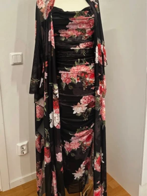 Svart blommig långklänning m - Supersnygg svart långklänning med rosa och röda blommor, figurnära passform och tunna axelband. Klänningen har rynkade detaljer och kommer med en matchande sjal i samma blommiga tyg. Perfekt för dig som vill sticka ut med en elegant look.
