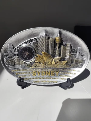 Souvenirplatta Sydney med klocka - Dekorativ oval souvenirplatta i silver- och guldfärgad metall med relief av Sydney skyline och text. Inbyggd liten klocka till vänster. Perfekt som prydnad eller samlarobjekt.