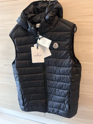 Svart dunväst från Moncler - Snygg svart dunväst från Moncler med huva och klassisk logotyp på bröstet. Västen har quiltad design, dragkedja framtill och är tillverkad i glansigt material. Perfekt för lager-på-lager och streetstyle.
