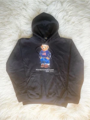 Svart hoodie Polo Bear Ralph Lauren - Svart hoodie från Ralph Lauren med ikoniska Polo Bear-trycket framtill. Hoodien har huva med dragsko och en stor magficka. Björnen på framsidan bär en blå tröja med amerikanska flaggan och jeans. Perfekt för en avslappnad och cool stil.