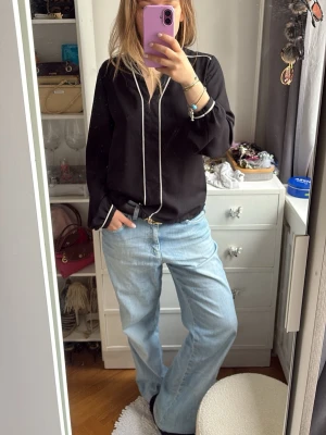 Svart blus med vita detaljer Atmosphere - Stilren svart blus från Atmosphere med vita kontrastlinjer längs v-ringningen och ärmsluten. Blusen har en avslappnad passform och långa ärmar, perfekt att matcha med jeans eller kjol. Materialet känns mjukt och följsamt.