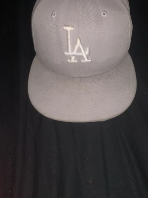 Grå LA 59FIFTY keps från New Era - Säljer en stilren grå keps från New Era, modell 59FIFTY, med LA-logga broderad framtill i vitt. Klassisk flat brim och ventilerande hål upptill. Perfekt för dig som gillar streetwear och sportig stil. Materialet är bomull och polyester.