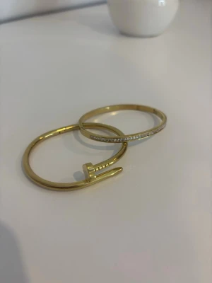 Två guldarmband med unik design - Säljer två snygga armband i guldfärg. Det ena har en cool spikformad design och det andra är ett klassiskt smalt armband med gnistrande stenar längs ovansidan. Super fina🤍