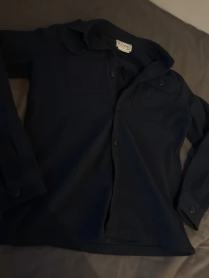 Mörkblå skjorta från Cope - Stilren mörkblå skjorta från Cope med klassisk krage, knäppning framtill och två bröstfickor med lock. Skjortan har långa ärmar med knappar vid manschetterna och är tillverkad i ett mjukt bomullstyg. Knappt använd