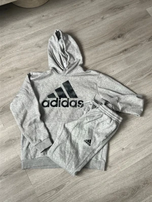 Grått Adidas loungewear set - Säljer ett grått loungewear set från Adidas med matchande hoodie och byxor. Hoodien har stor svart Adidas-logga på bröstet och byxorna har en mindre logga vid benet. Mjukt material och avslappnad passform, perfekt för chill dagar hemma.