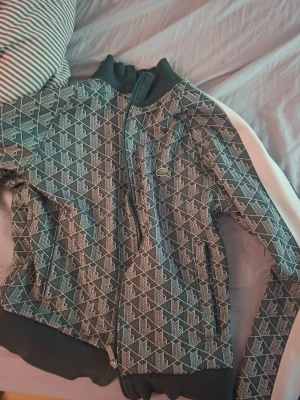 Mönstrad zip-jacka från Lacoste - Snygg jacka från Lacoste med grafiskt mönster i mörkgrönt och vitt. Jackan har hög krage, dragkedja framtill och ribbade muddar. Ikoniska Lacoste-loggan på bröstet. Perfekt för dig som vill sticka ut med stil.