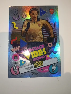 Match Attax Thierry Henry Arsenal-kort - Samlarkort från Topps Match Attax med Arsenal-legenden Thierry Henry. Kortet har holografisk yta, färgglada retrodetaljer och texten 'Vintage Vibes Legend'. Framsidan visar Henry i Arsenal-tröja och baksidan har Arsenal FC-logga. Perfekt för fotbollsfans och samlare.