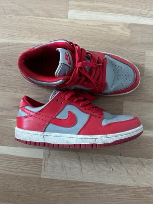 Nike Dunk Low röd och grå sneaker - Nike Dunk Low sneakers i rött och grått med klassisk Swoosh-logga på sidan. Ovandel i en mix av läder och textil, röda skosnören och yttersula. Perforerad tåbox för extra ventilation och vadderad plös med Nike-logga.