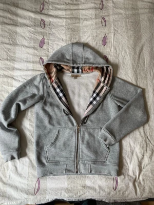 Grå hoodie från Burberry med dragkedja - Säljer en grå hoodie från Burberry med dragkedja och klassiskt rutigt foder i huvan. Hoodien har fickor framtill, ribbade muddar och en broderad Burberry-logga på bröstet. Perfekt för dig som gillar stilrena plagg med exklusiva detaljer. Skriv vid minsta funderingar😁