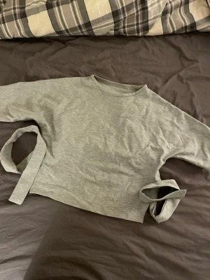 Grå croppad topp med knytband - Säljer en ljusgrå croppad topp med rund hals och korta ärmar. Toppen har snygga knytband i sidorna som ger en unik look. Perfekt till jeans eller kjol för en chill och trendig stil.
