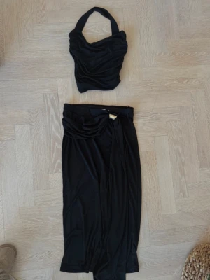 Svart croppad halterneck topp + kjol - Trendig svart croppad topp med halterneck och draperad design. Toppens material är mjukt och följsamt, perfekt för en stilren look. Matchande  kjol till helt nytt 