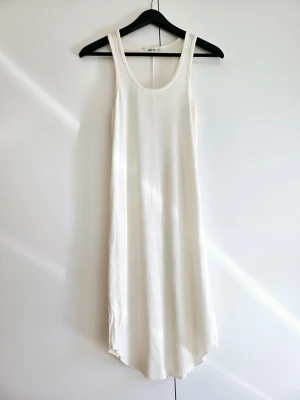 Vit ribbad klänning i bomull - Snygg vit midi klänning i ribbad bomull med ärmlös design och rund halsringning. Perfekt för varma dagar och enkel att styla med accessoarer. Klänningen har en rak kropsformad passform och mjukt material som känns skönt mot huden.