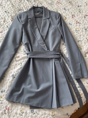 Grå blazerklänning från Zara - Snygg grå blazerklänning från Zara med omlott-design och bälte i midjan. Klänningen har lång ärm, klassisk krage och stilren look. Perfekt för dig som gillar en trendig och sofistikerad stil.