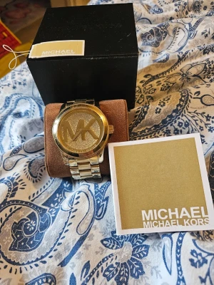 Michael Kors Klocka  - Klocka från Michael Kors modellen är: MK5706 Klockan har ett armband i rostfritt stål och i färgen roséguld samt en urtavla i färgen guld fylld med glittriga kristaller. Klockarmbandets bredd: 24mm  Vattentåligt djup: 100m/330fot/10ATM  Klockans urverk: Kvarts  Vid köp följer original kartong, info bok samt prislapp etiketten (allt syns på bild)  Fungerar felfritt har fått en ny klocka så denna används ej längre därav säljs den.