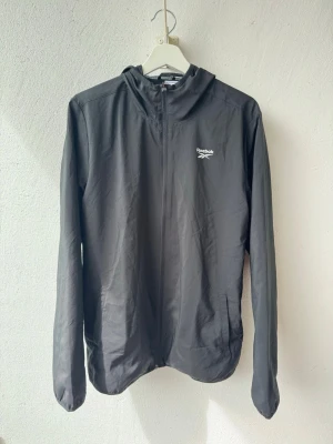 Svart vindjacka från Reebok med huva - Snygg svart vindjacka från Reebok med dragkedja framtill och praktisk huva. Jackan har två sidofickor och Reebok-logga på bröstet. Tillverkad i lätt och vindtätt material som passar perfekt för blåsiga dagar. 1 gång använd 9.9/10