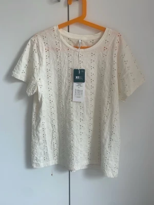 Vit broderad t-shirt från ONLY - Snygg vit t-shirt från ONLY med broderade detaljer och små hålmönster. T-shirten är kortärmad och gjord i mjuk bomull, perfekt för en fräsch och stilren look. Passar till både jeans och kjol för en trendig outfit.