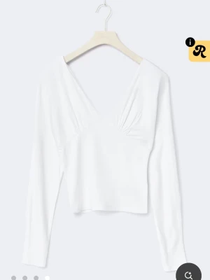 Vit omlottopp med v-ringning - Snygg vit omlottopp med djup v-ringning och långa ärmar. Toppen har en figurnära passform och lätt rynkade detaljer vid bysten. Perfekt att styla med jeans eller kjol för en trendig look.