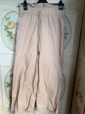 Beiga linnebyxor från Noisy May - Snygga beiga linnebyxor från Noisy May med hög midja och resår samt dragsko. Vida ben och avslappnad passform, perfekta för en chill och trendig look. Tillverkade i ett lätt och luftigt material som passar bra till varmare dagar.
