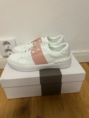 Valentino vita sneakers med rosa detalj - Snygga vita sneakers från Valentino med en bred rosa rem över vristen och ikoniska nitar längs sulan. Skorna har en chunky sula och är tillverkade i läder med vita skosnören. Perfekta för dig som vill sticka ut med en stilren men unik look.