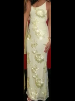 Lång vit klänning med blommigt mönster - Elegant långklänning i gul nyans med stora blommiga mönster i ljusgult. Klänningen har ärmlös design och smala axelband, samt en figurnära passform. Perfekt för speciella tillfällen när du vill sticka ut med en stilren look.