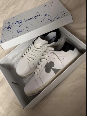 Vita sneakers från The Gallerist - Snygga vita sneakers från The Gallerist med svart häl och en grå klubb-symbol på sidan. Skorna har klassisk låg profil, vita snören och är gjorda i läder. Perfekta för dig som vill ha en stilren men unik look. Passar allt från storlek 42-43. Typ helt nya, (använda 4-3 gånger)