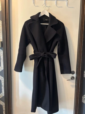 Svart lång kappa med knytskärp - Stilren svart kappa med lång passform och brett slag. Kappan har ett knytskärp i midjan och långa ärmar. Perfekt för kyliga dagar och ger en klassisk look. Materialet ser ut att vara ullblandning för extra värme.