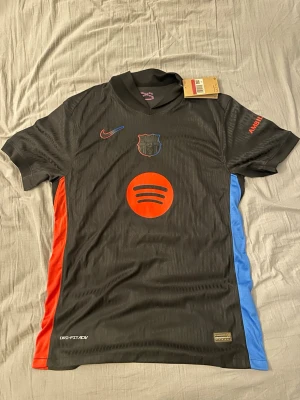 FC Barcelona svart matchtröja Nike - Snygg svart FC Barcelona matchtröja från Nike med röda och blå detaljer på sidorna. Tröjan har klubbmärke och Nike-logga på bröstet, samt Spotify-logga i rött framtill. Tillverkad i lätt och ventilerande material, perfekt för fotbollsträning eller match.