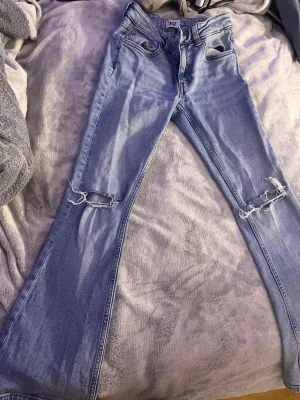Ljusblå bootcut jeans med slitningar - Snygga ljusblå bootcut jeans med slitna hål vid knäna. Jeansen har klassisk femficksdesign och dragkedja med knapp. Perfekta för en trendig och avslappnad stil. Materialet är denim i bomull.