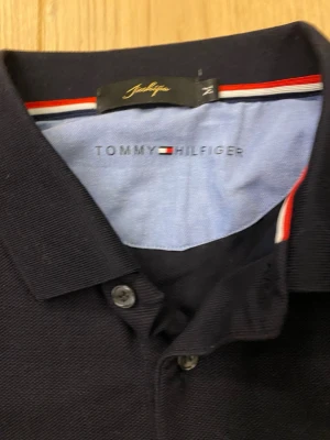 Svart pikétröja från Tommy Hilfiger - Klassisk svart pikétröja från Tommy Hilfiger med två knappar framtill och diskret logga broderad på bröstet. Tröjan har krage och är tillverkad i ett bekvämt bomullsmaterial. Perfekt för en stilren och avslappnad look.