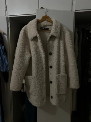 Krämvit teddyjacka från Zara - Mysig krämvit teddyjacka från Zara med stora framfickor och klassisk krage. Jackan har knäppning med svarta knappar och är tillverkad i ett fluffigt teddy-material som håller dig varm. Perfekt för kyliga dagar och enkel att styla till olika outfits.