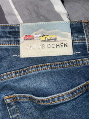 Blå jeans från Jacob Cohën - Snygga blå jeans från Jacob Cohën med klassisk femficksdesign och kontrastsömmar. Jeansen har en cool patch med bilar och berg på bakfickan. Finns digitalt kvitto från Nk det är bara att fråga i dm.