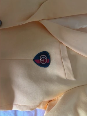 Gul kavaj från Gucci med emblem - Snygg gul kavaj från Gucci med klassiskt GG-emblem på bröstfickan. Kavajen har en stilren design med mjukt tyg och innerfoder. Märket syns tydligt på insidan och detaljerna har en lyxig känsla. Perfekt för dig som vill sticka ut med något unikt och exklusivt.