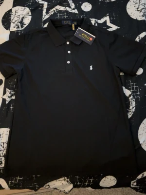 Svart pikétröja från Polo Ralph Lauren - Klassisk svart pikétröja från Polo Ralph Lauren med vit broderad logga på bröstet. Tröjan har korta ärmar, krage och tre knappar framtill. Perfekt för en stilren och avslappnad look.