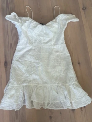 Vit spetsklänning med volang - Söt vit klänning med tunna axelband och offshoulder-detalj. Klänningen har broderad spets över hela och en volang längst ner för extra flow. Perfekt för sommarens alla tillfällen.
