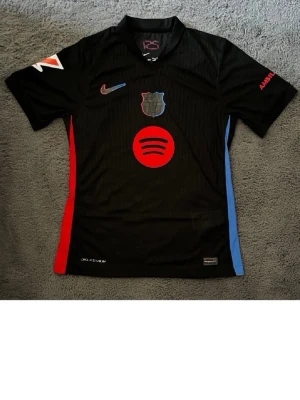 Svart FC Barcelona matchtröja Nike - Snygg svart FC Barcelona matchtröja från Nike med röda och blå detaljer på sidorna. Tryck med Spotify-logga framtill och Lamine Yamal 19 på ryggen. Klubbmärke och Nike-logga på bröstet, kort ärm och sportig passform. Perfekt för fotbollsträning eller support.