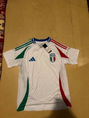 Italien Adidas fotbollströja vit - Snygg vit fotbollströja från Adidas med Italiens landslagsmärke på bröstet. Tröjan har blå krage, gröna och röda detaljer på axlar och sidor samt Adidas logga. Tillverkad i lätt och ventilerande material, perfekt för träning eller match.